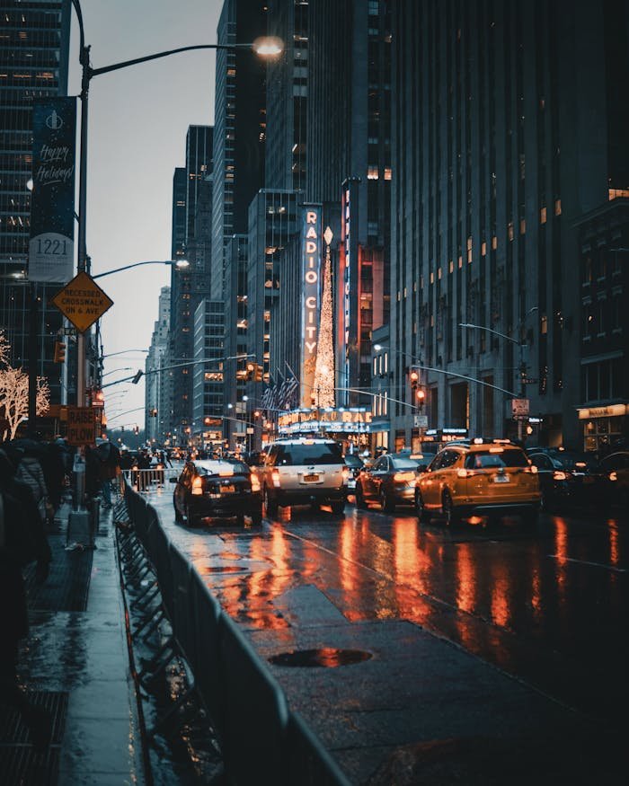 hero-img-01 Dramatic city street view of New York's Radio City at night with reflections and taxis.