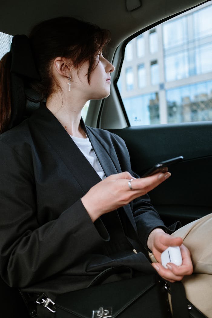 our-services-1 A woman with a ponytail and earrings uses a smartphone in the back of a car, focusing outside.